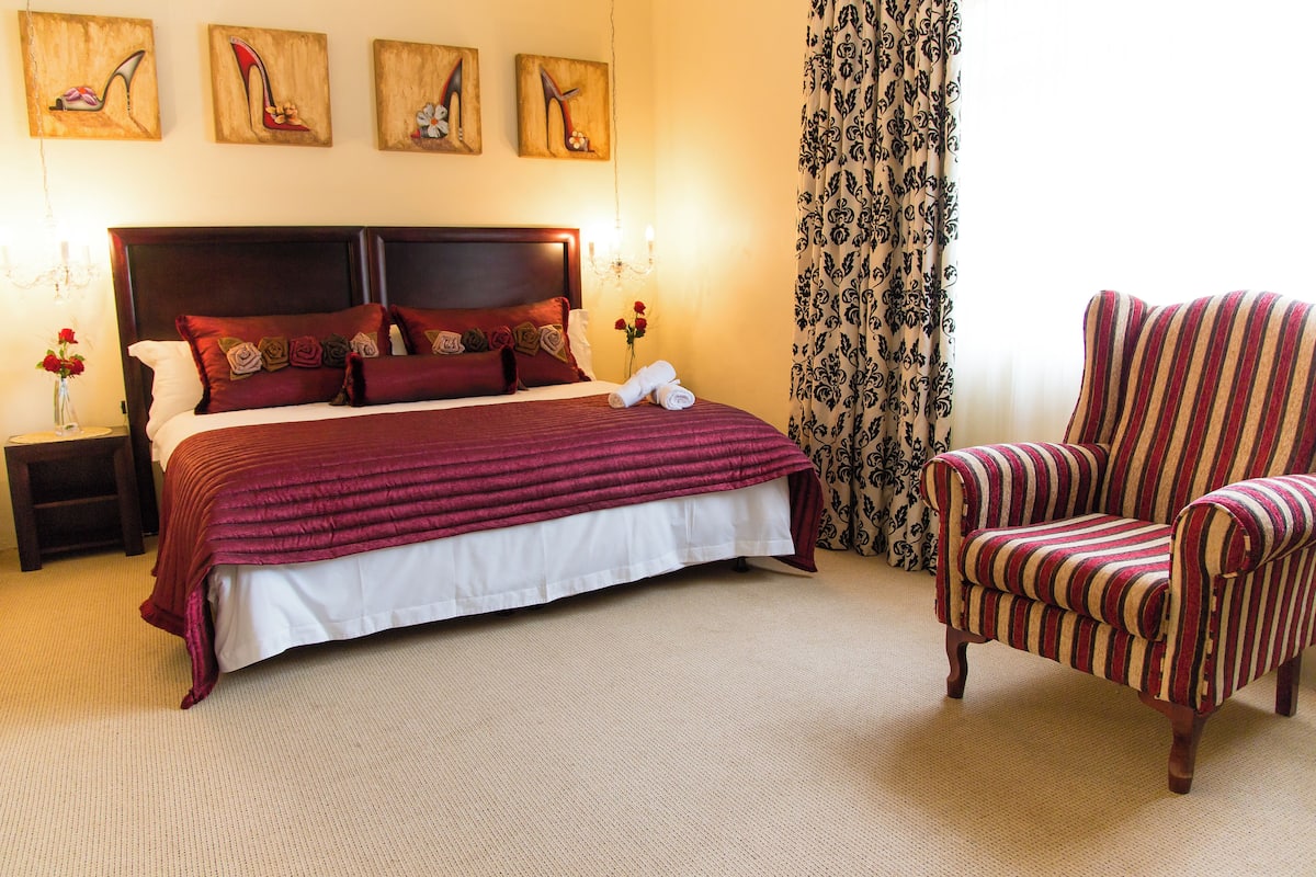A neatly made bed with a red and white bedspread, a striped armchair, a nightstand with a vase of flowers, and three paintings on the wall.