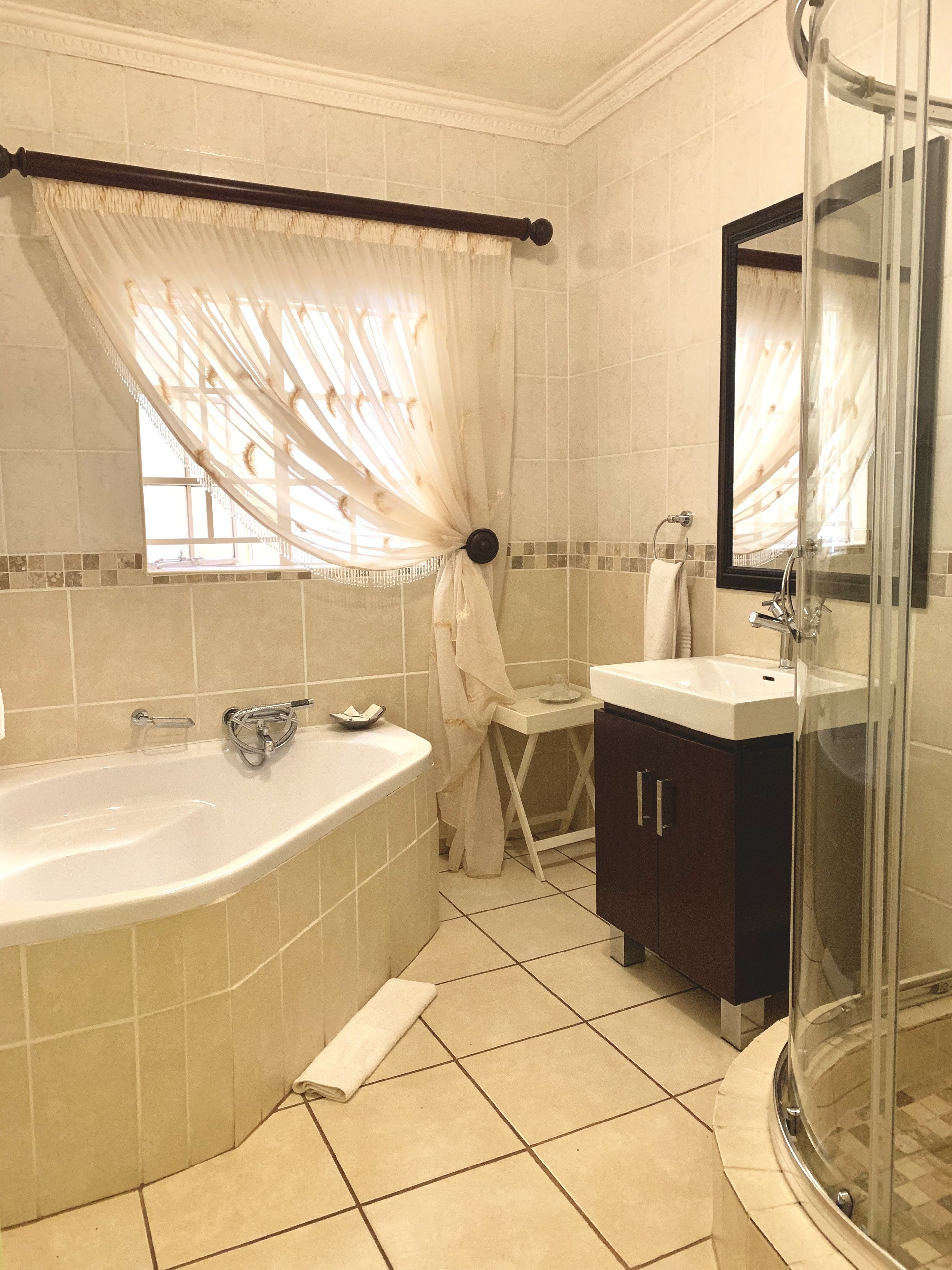 A bathroom with a bathtub, a glass-enclosed shower, a vanity with a mirror, and a window with sheer curtains.