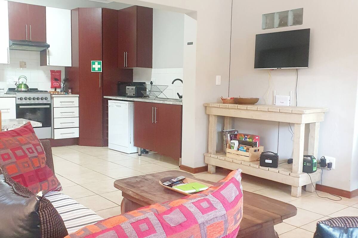 A living room with a sofa, coffee table, and a television. There is a kitchen in the background with cabinets and appliances.