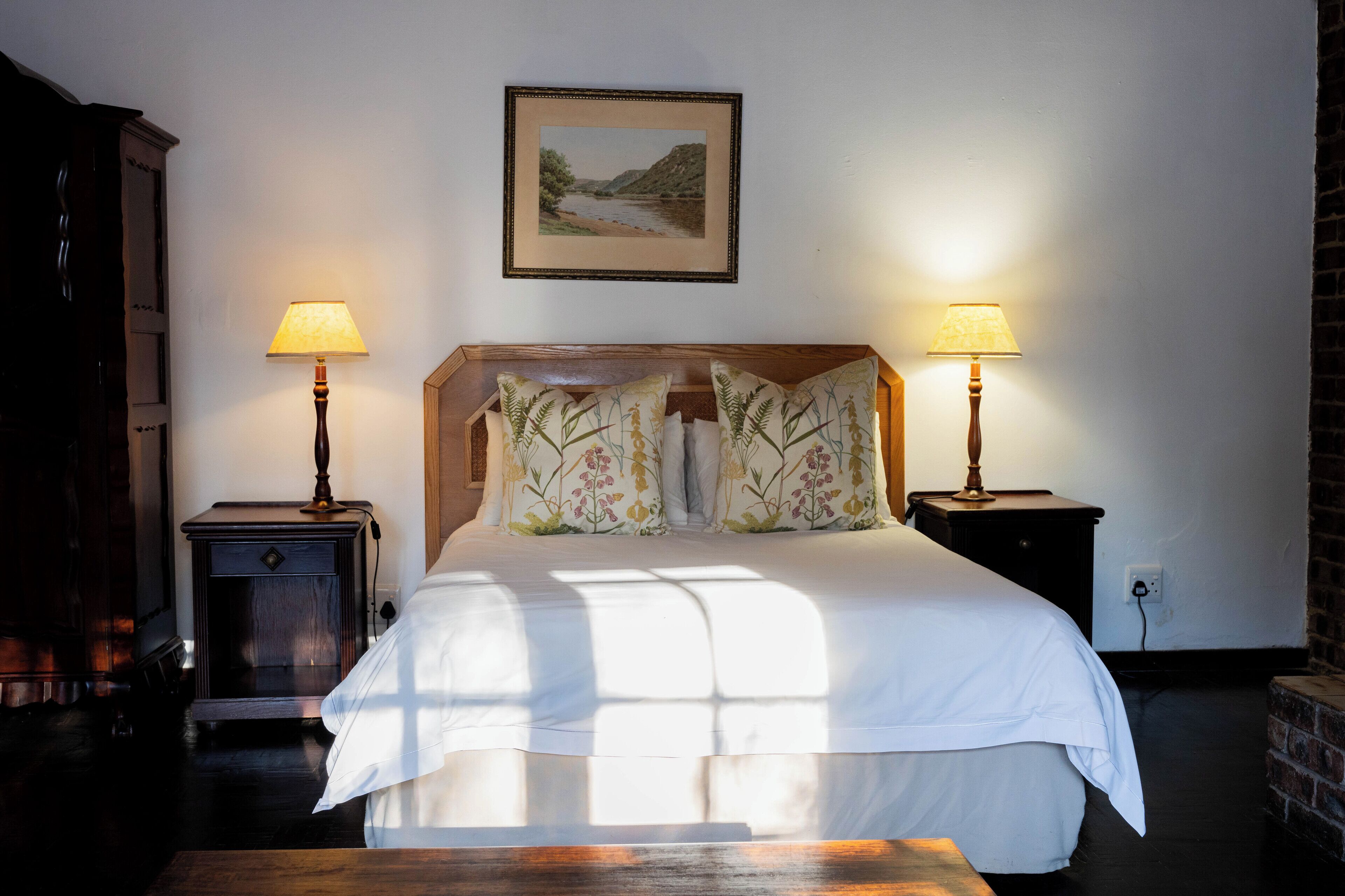 Una cama bien hecha con almohadas florales, dos lámparas de noche y un cuadro con un paisaje en la pared.