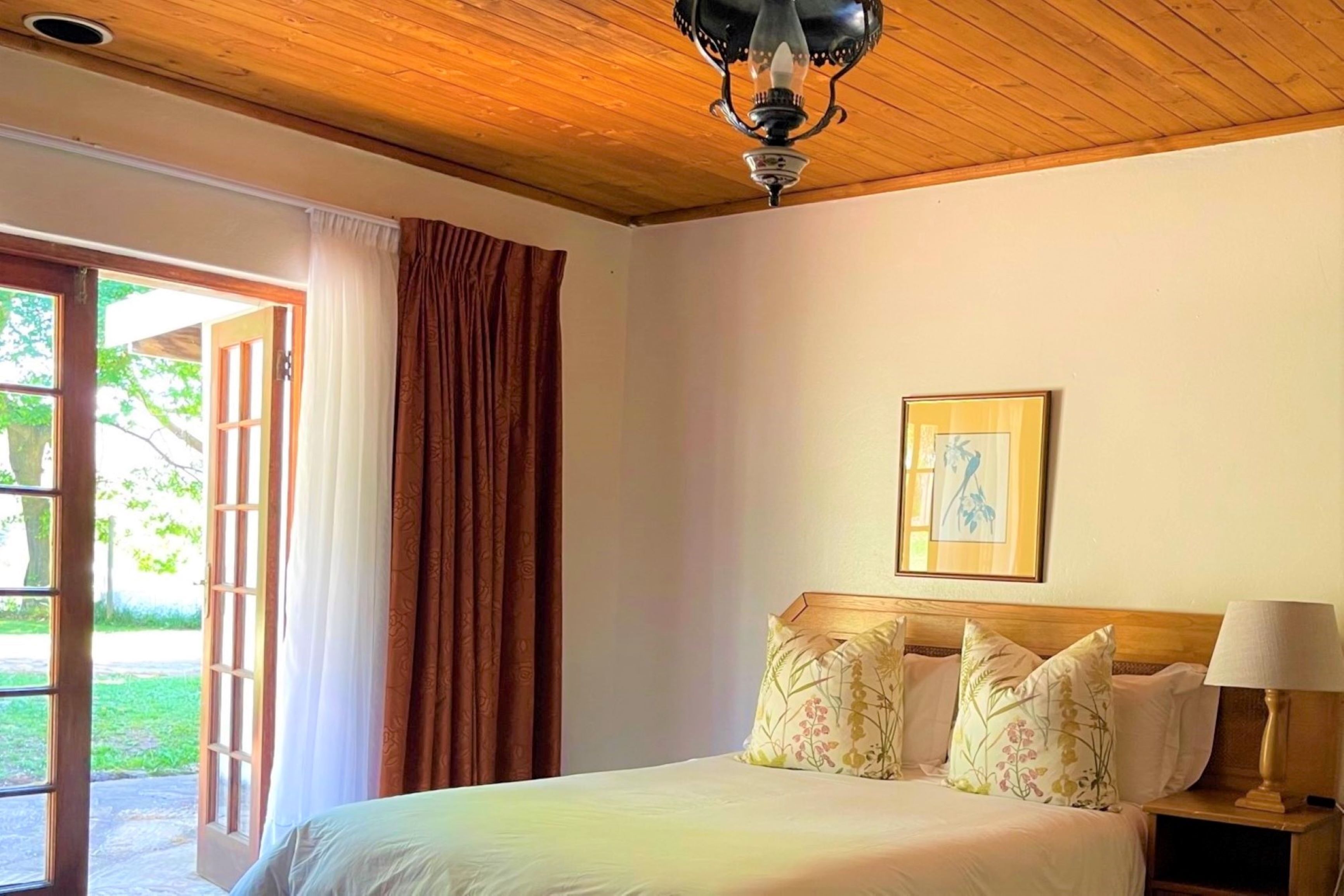 A bedroom with a wooden ceiling, a bed with floral pillows, a nightstand with a lamp, and a framed picture on the wall.