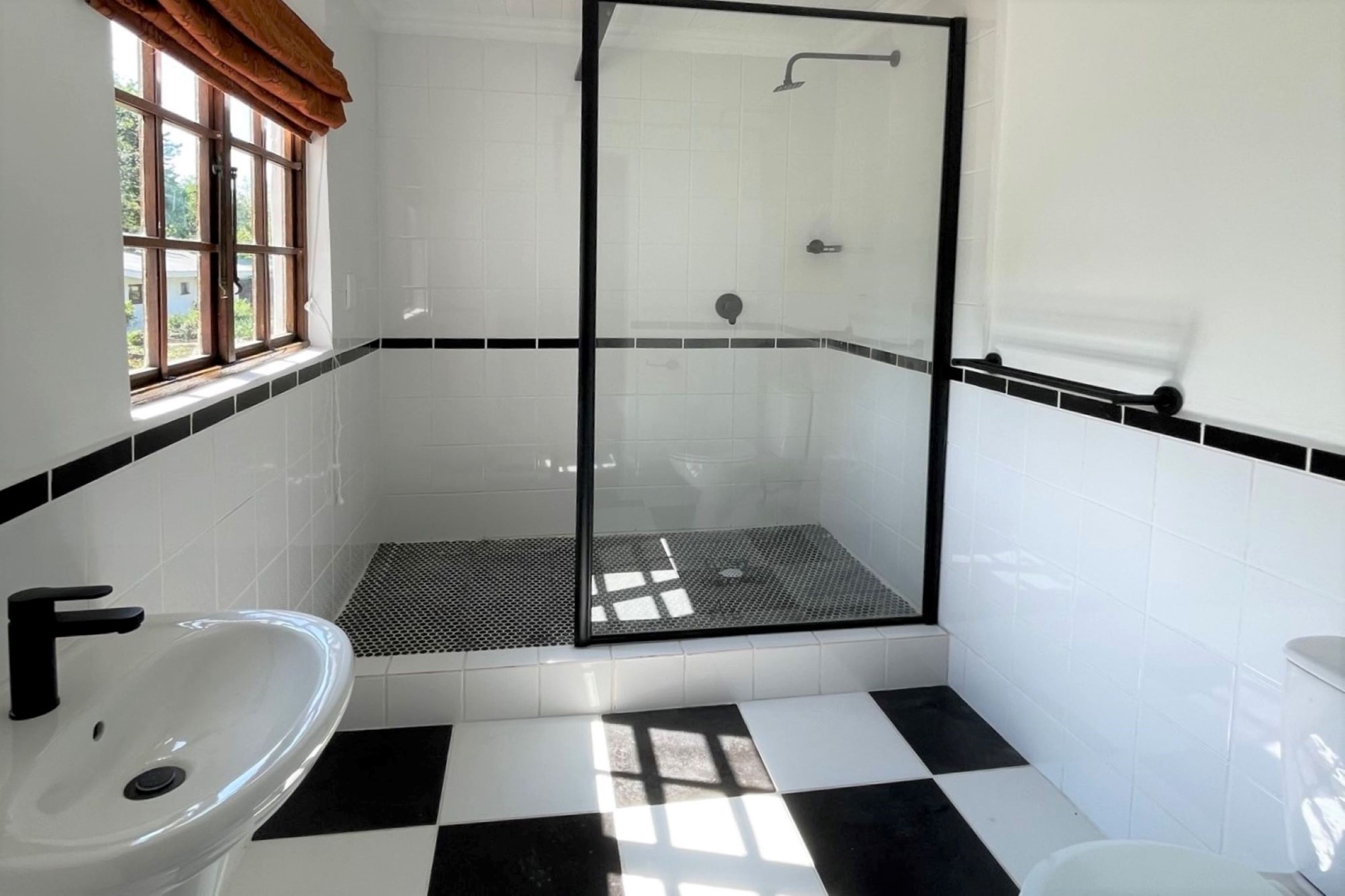 A bathroom with a black and white checkered floor, a glass-enclosed shower, and a window with a brown valance.