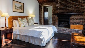Quarto com cama grande, cabeceira de madeira, duas mesas de cabeceira com luminárias, lareira de tijolos e um quadro na parede.