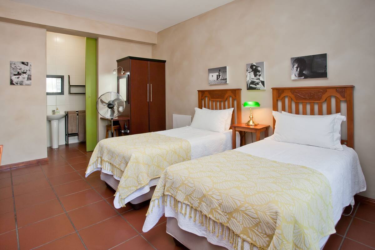 Une chambre d’hôtel avec deux lits, un mur vert, une armoire en bois, un bureau avec une chaise et un ventilateur.
