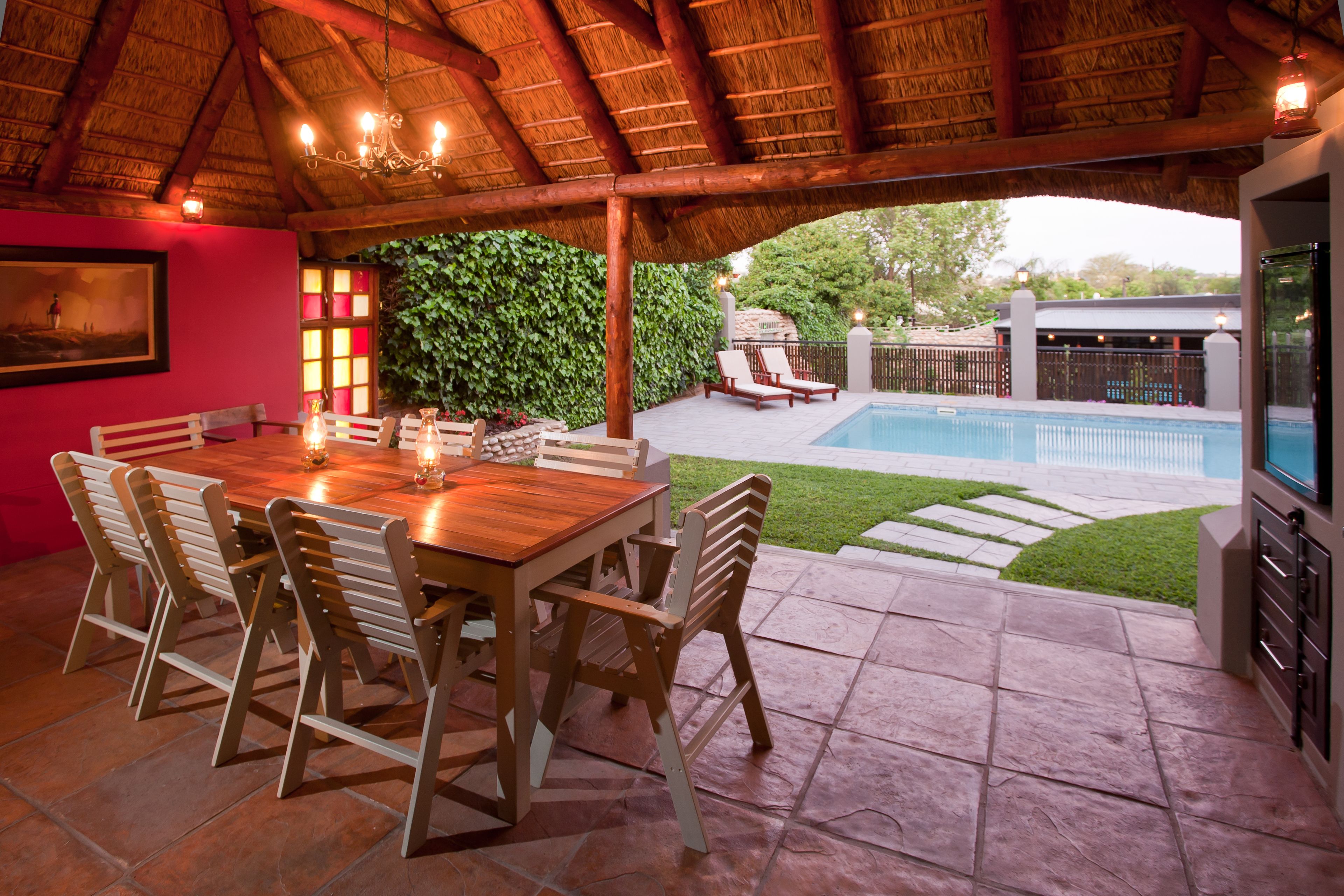 An outdoor dining area with a wooden table and chairs, a thatched roof, and a pool in the background.