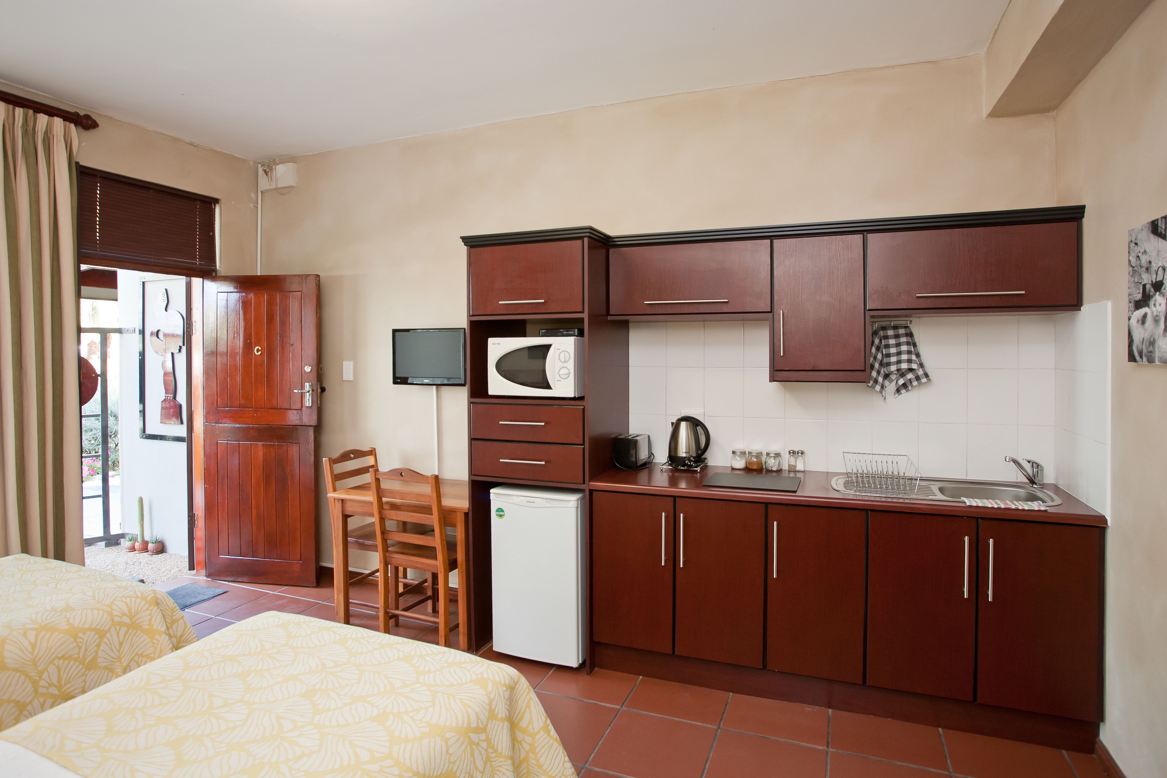 A compact hotel room with a kitchenette, a bed with patterned bedding, a small refrigerator, and a wooden chair.