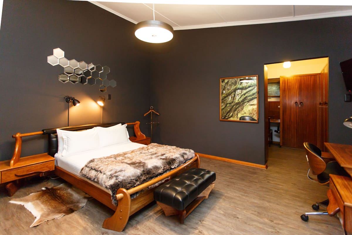 Une chambre avec un lit en bois, un banc, un bureau et une chaise. On y trouve également un porte-manteau et un tableau accroché au mur.