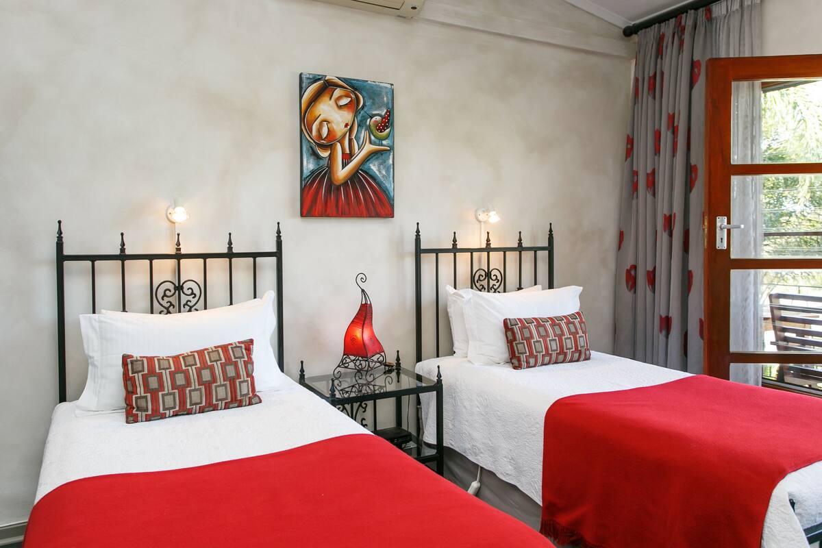 Une chambre d’hôtel avec deux lits, chacun recouvert d’une couverture rouge et agrémenté d’oreillers à motifs. Un tableau orne le mur, une petite table avec une lampe rouge s’y trouve, et une fenêtre est habillée de rideaux.