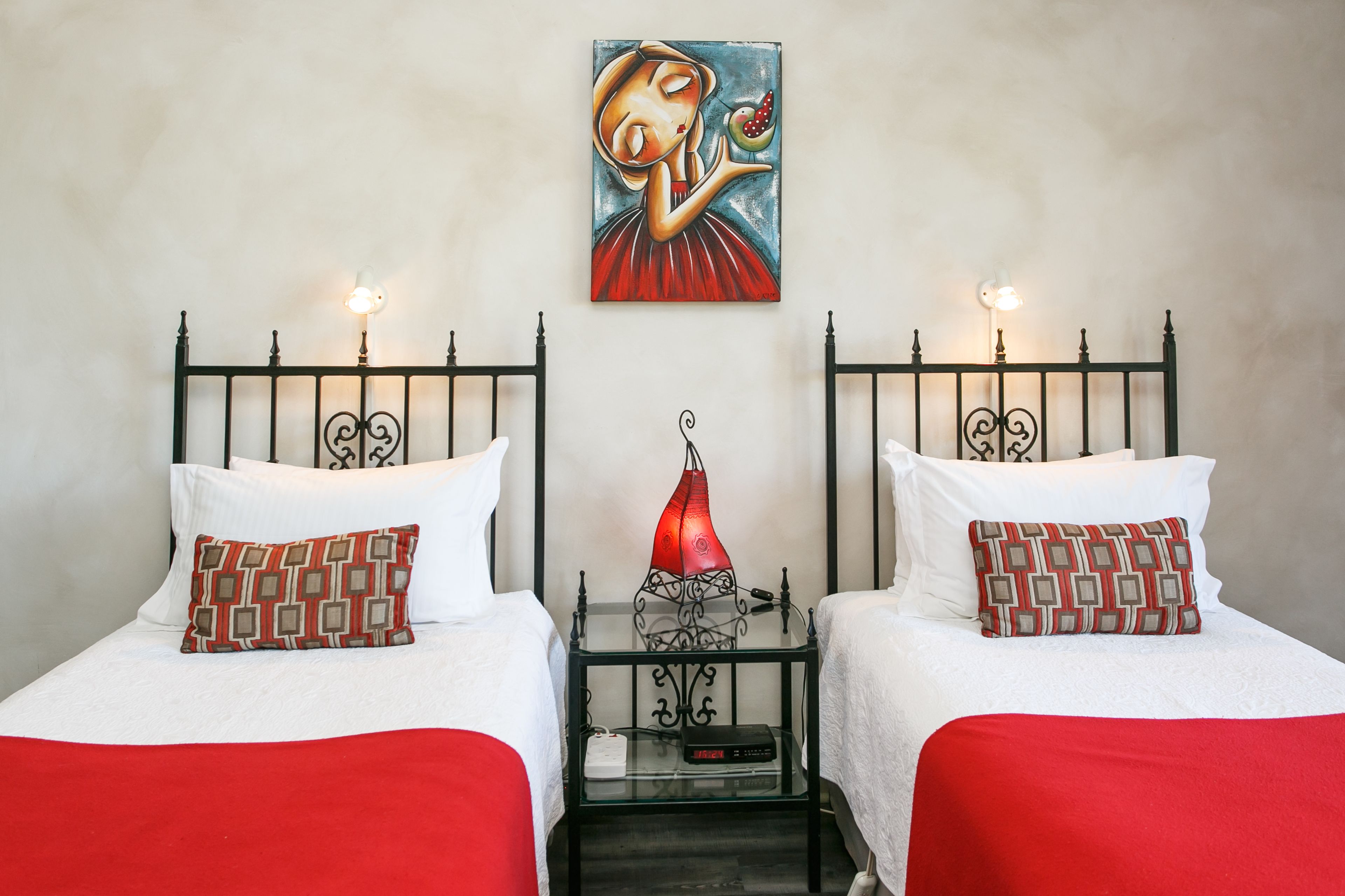 Una habitación de hotel con dos camas, cada una con una almohada estampada en rojo y blanco, una mesita de noche con lámpara roja, y un cuadro en la pared.