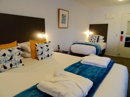 Habitación de hotel con dos camas, cada una con ropa de cama blanca y mantas azules. Hay almohadones decorativos, un cuadro enmarcado en la pared y una mesita de noche con un teléfono y gafas.
