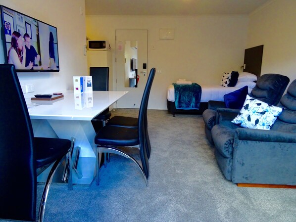 A hotel room with a dining table, chairs, a sofa, a bed with a blanket, and a television displaying a show. - BKs Premier Motel Hamilton (Hamilton)