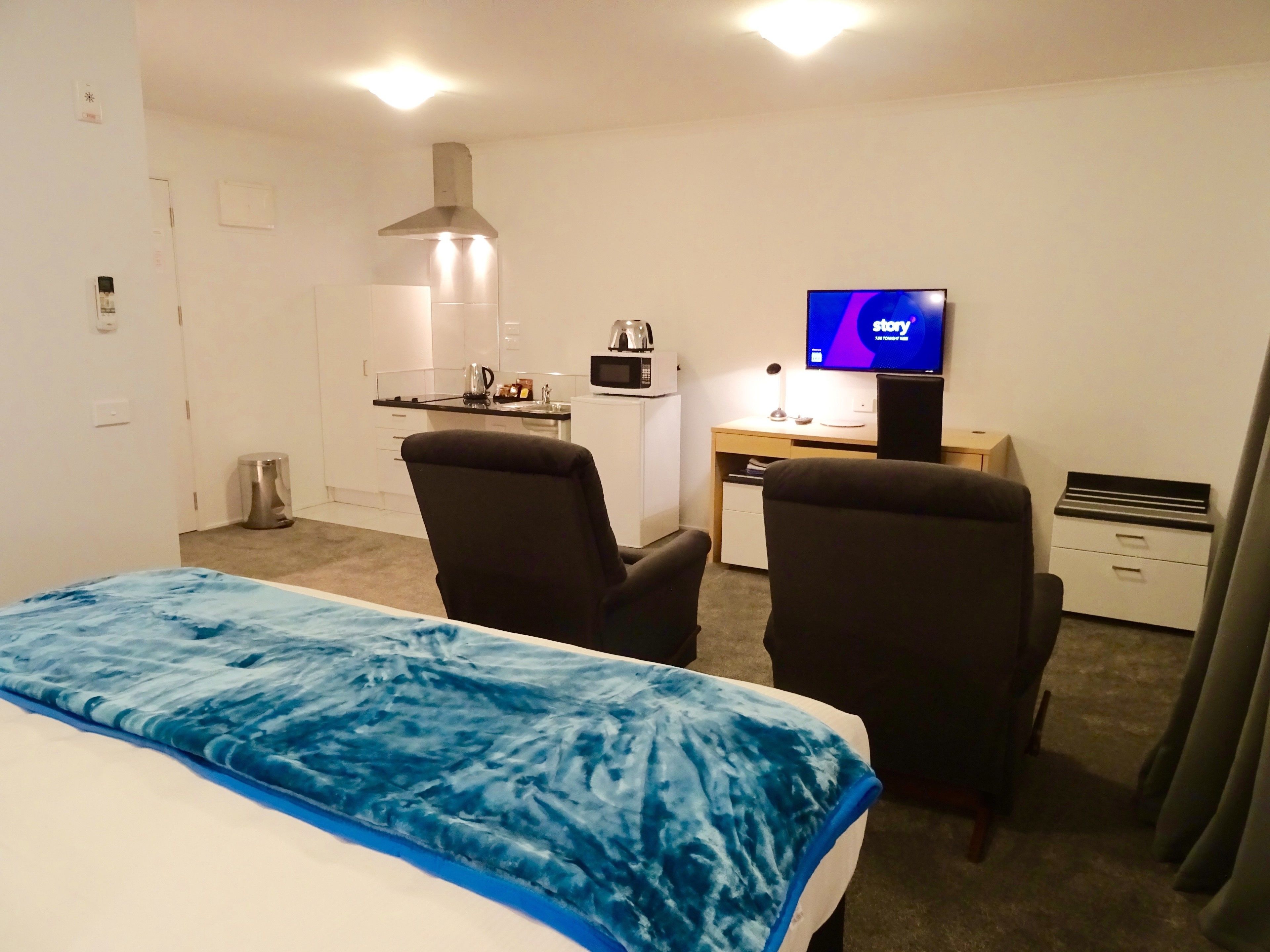 Habitación de hotel con una cama, dos sillones, un escritorio con computadora y televisor, microondas y kitchenette.