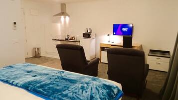 Habitación de hotel con una cama, dos sillones, un escritorio con computadora y televisor, microondas y kitchenette.