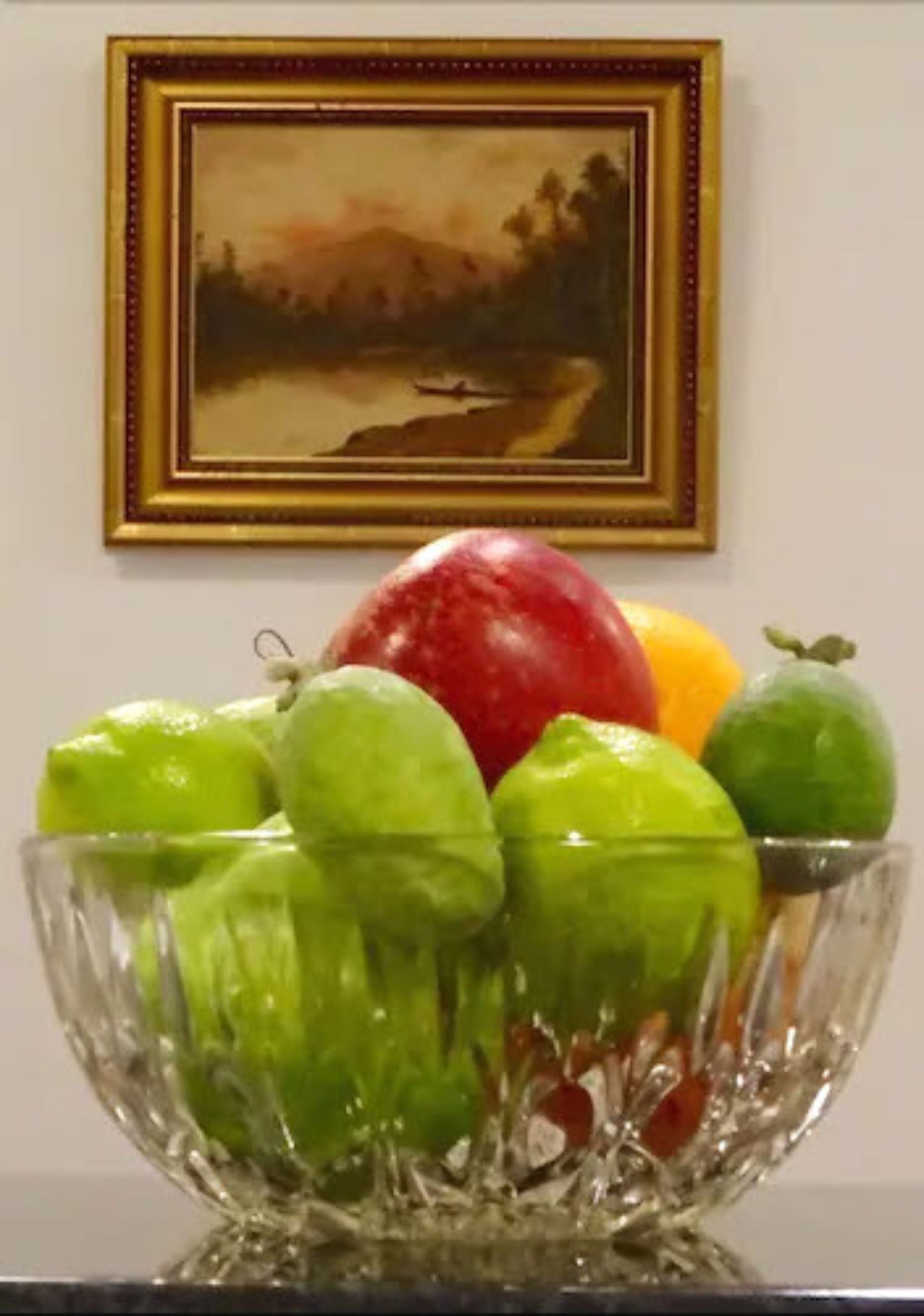A glass bowl with green apples and a red apple on a table.
