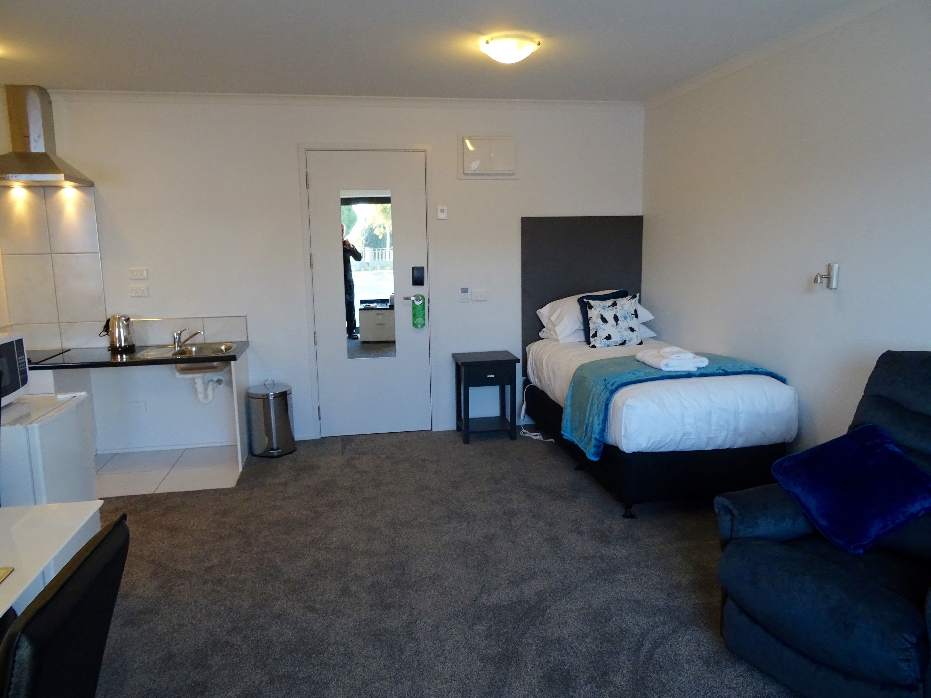 A compact hotel room with a kitchenette, a bed with a blue blanket, a dark armchair, and a small bedside table.