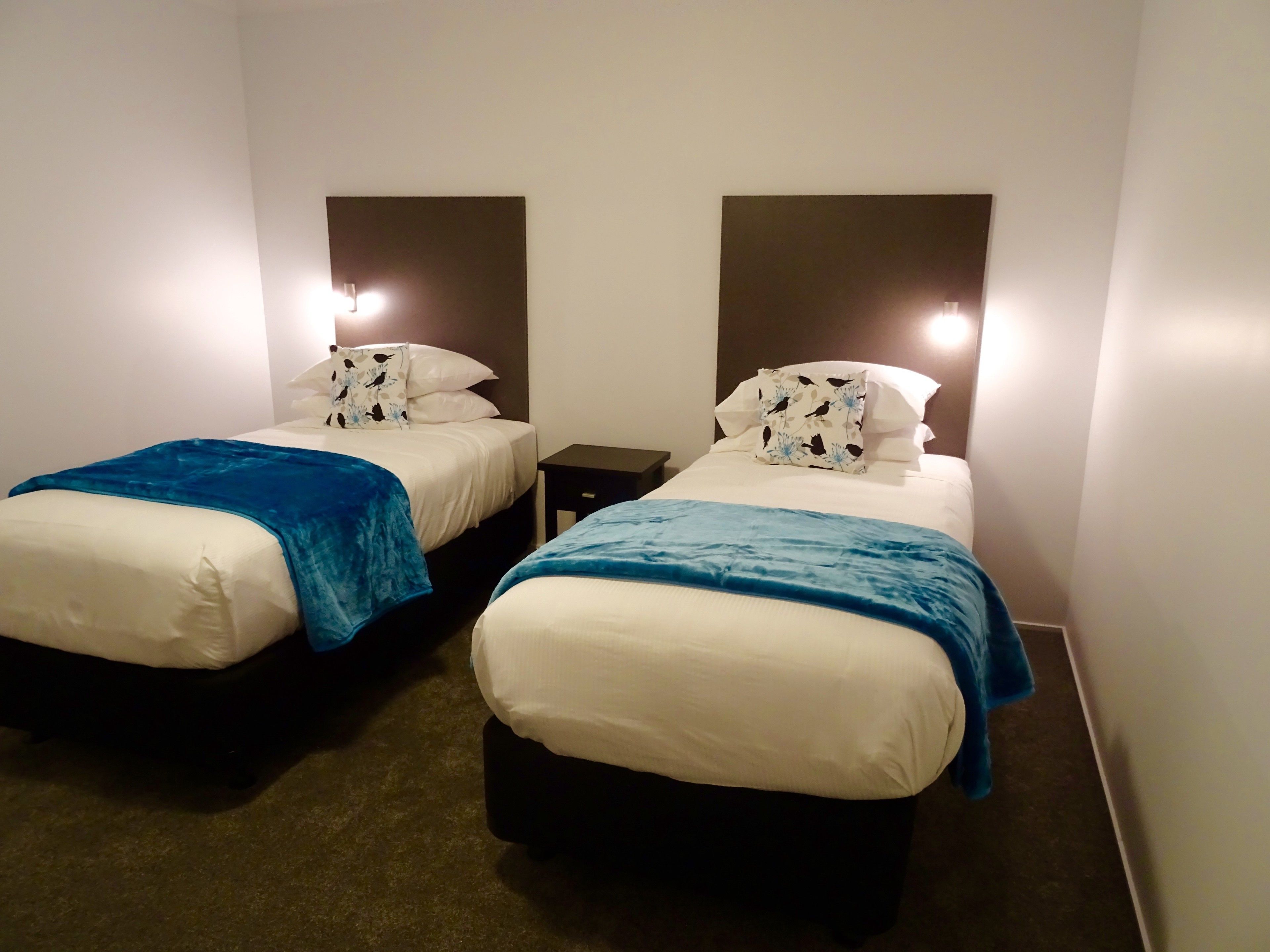 A hotel room with two beds, each with a blue blanket and white bedding. There are bedside tables with lamps.