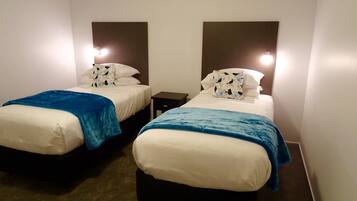 A hotel room with two beds, each with a blue blanket and white bedding. There are bedside tables with lamps.