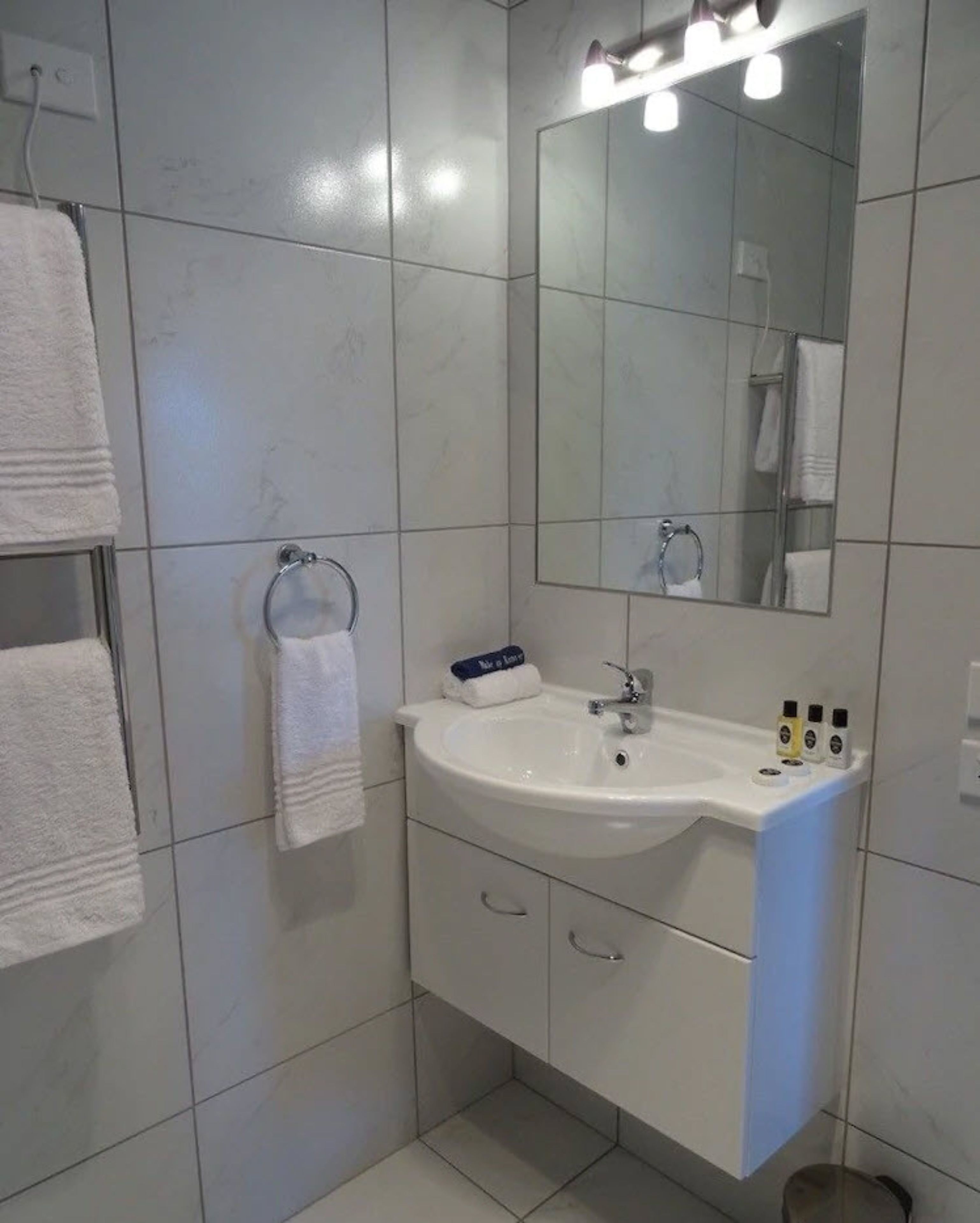 A modern bathroom with a white sink, mirror, and tiled walls. There are towels hanging on a rack and a small shelf with toiletries.