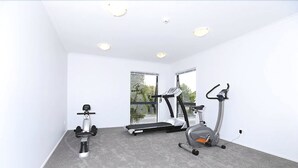 Quarto com esteira ergométrica, elíptico e bicicleta fixa, piso coberto de carpete, paredes brancas e grandes janelas.
