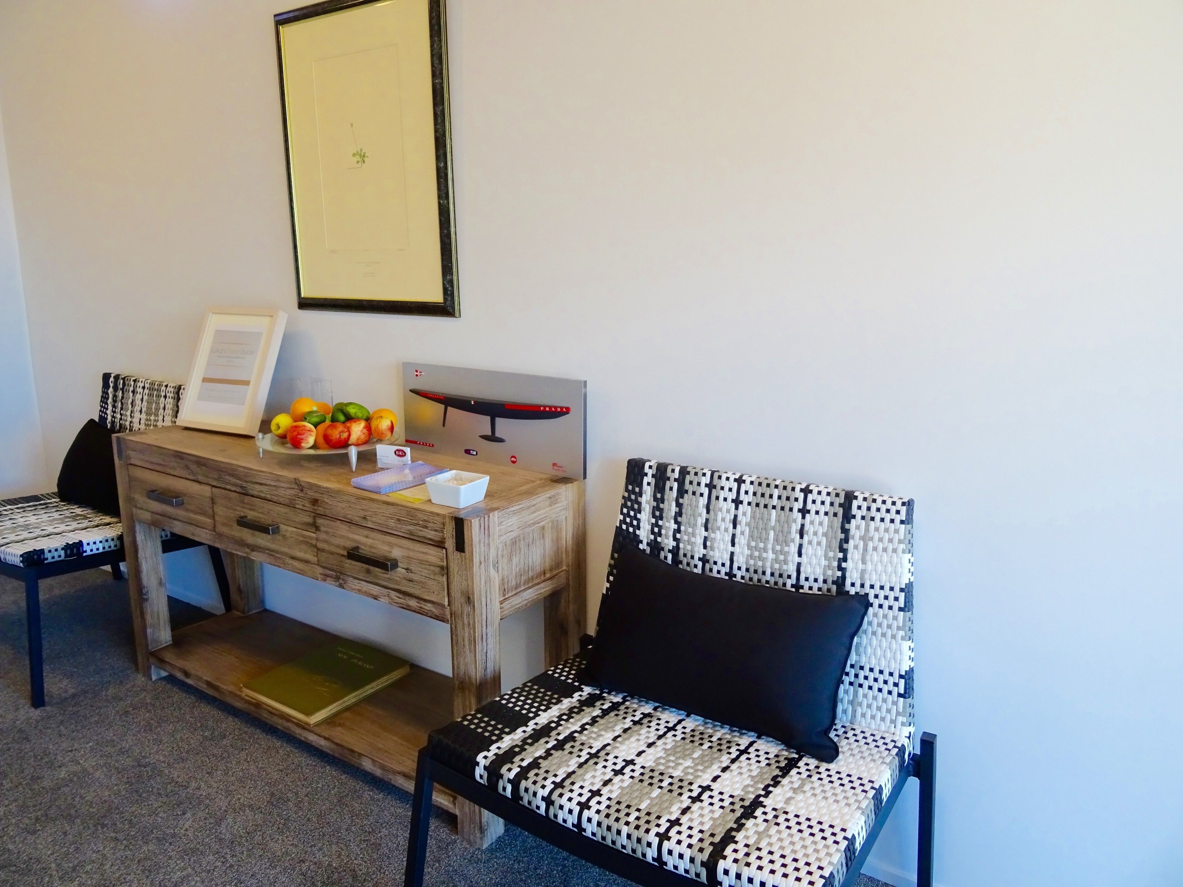 A room with a wooden desk, a framed certificate, a fruit bowl, a chair with a patterned cushion, and a wall-mounted picture.