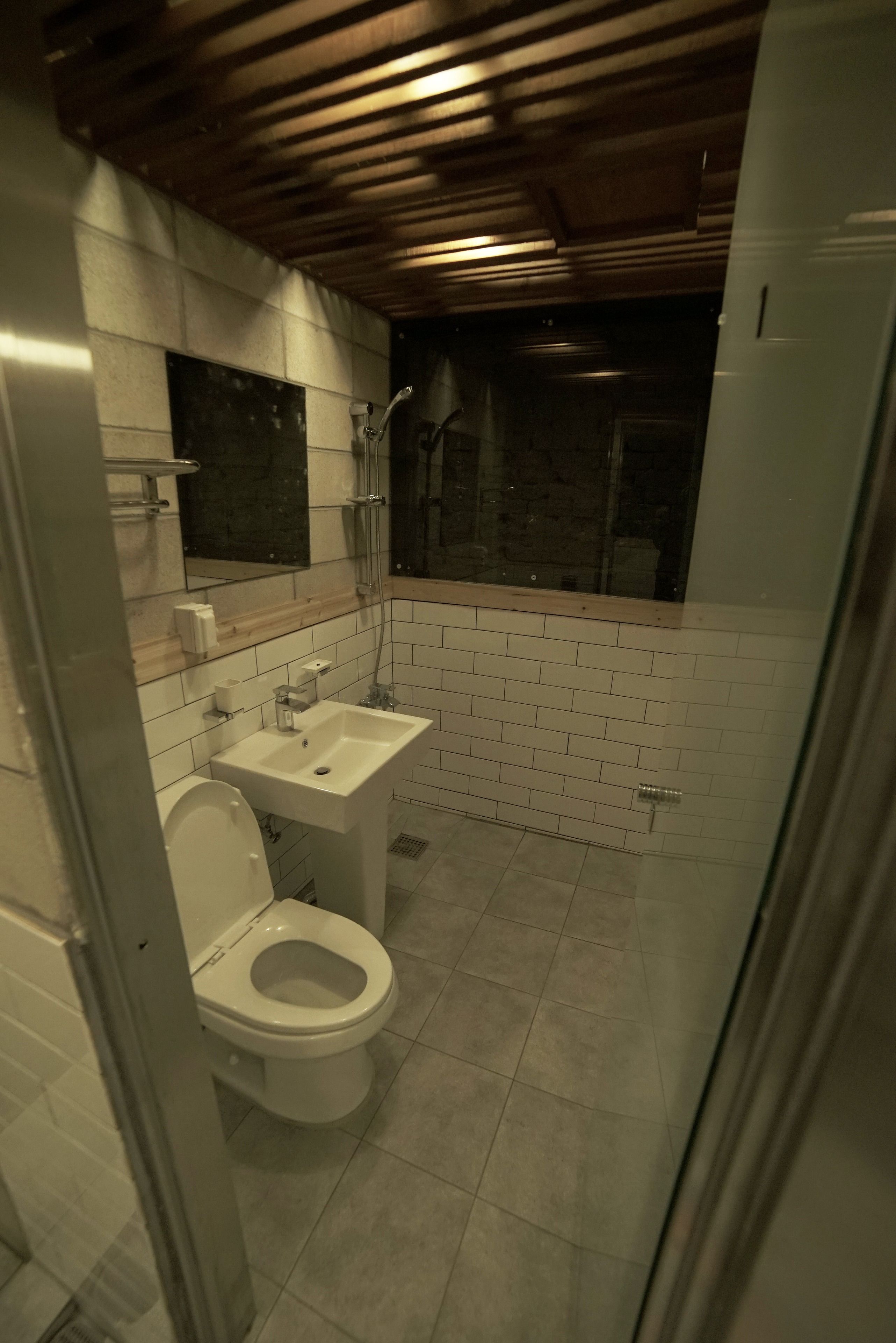 A small, compact bathroom with a white toilet, a sink, and a shower. The walls are tiled, and there is a wooden ceiling.