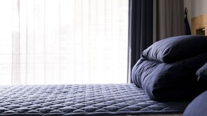 Un lit avec une couverture matelassée et des oreillers, une tête de lit en bois et des rideaux légers qui laissent passer la lumière naturelle.