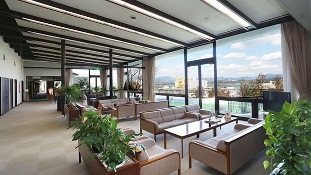 A spacious lounge area with sofas and a coffee table, large windows offering a view of the city, and a ceiling with linear lighting fixtures. - Hamayu Kuroshio Sanso (Shirahama)