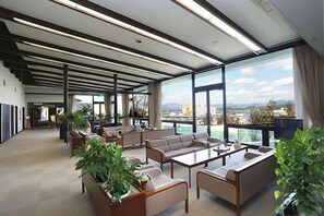 A spacious lounge area with sofas and a coffee table, large windows offering a view of the city, and a ceiling with linear lighting fixtures. - Hamayu Kuroshio Sanso (Shirahama)
