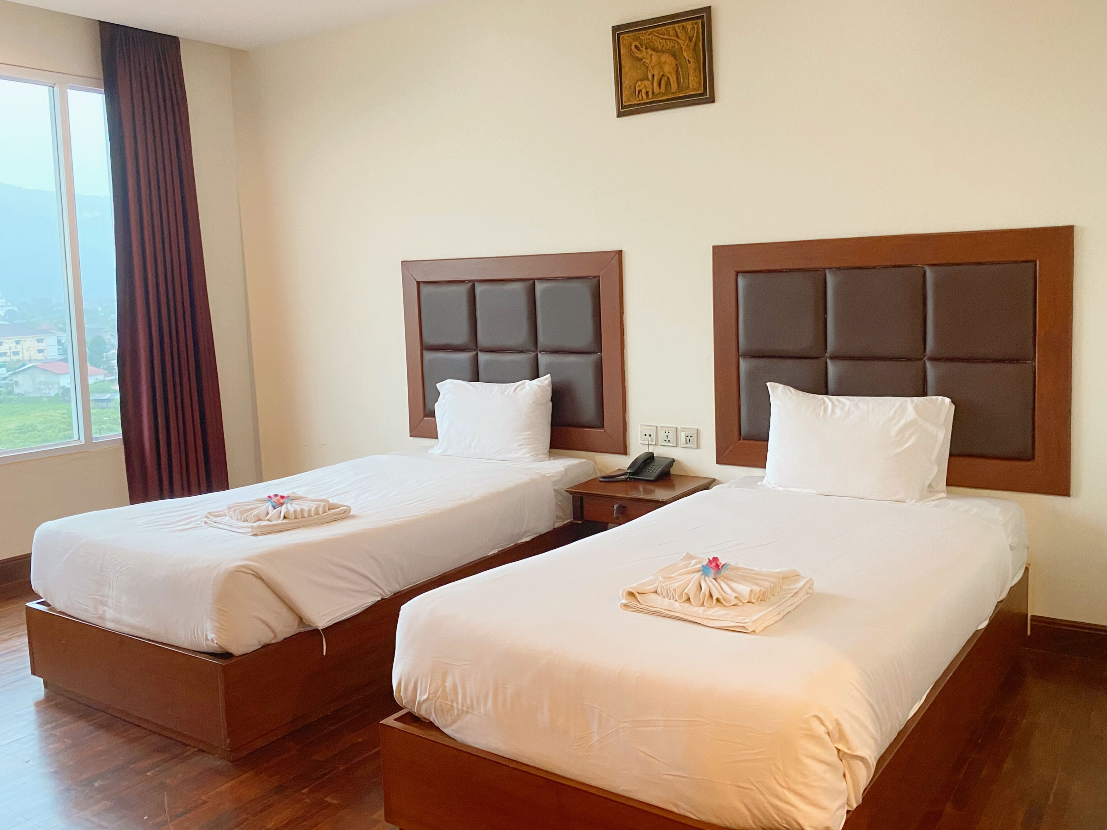 A hotel room with two single beds, wooden headboards, white bedding, a bedside telephone, and a window with curtains.