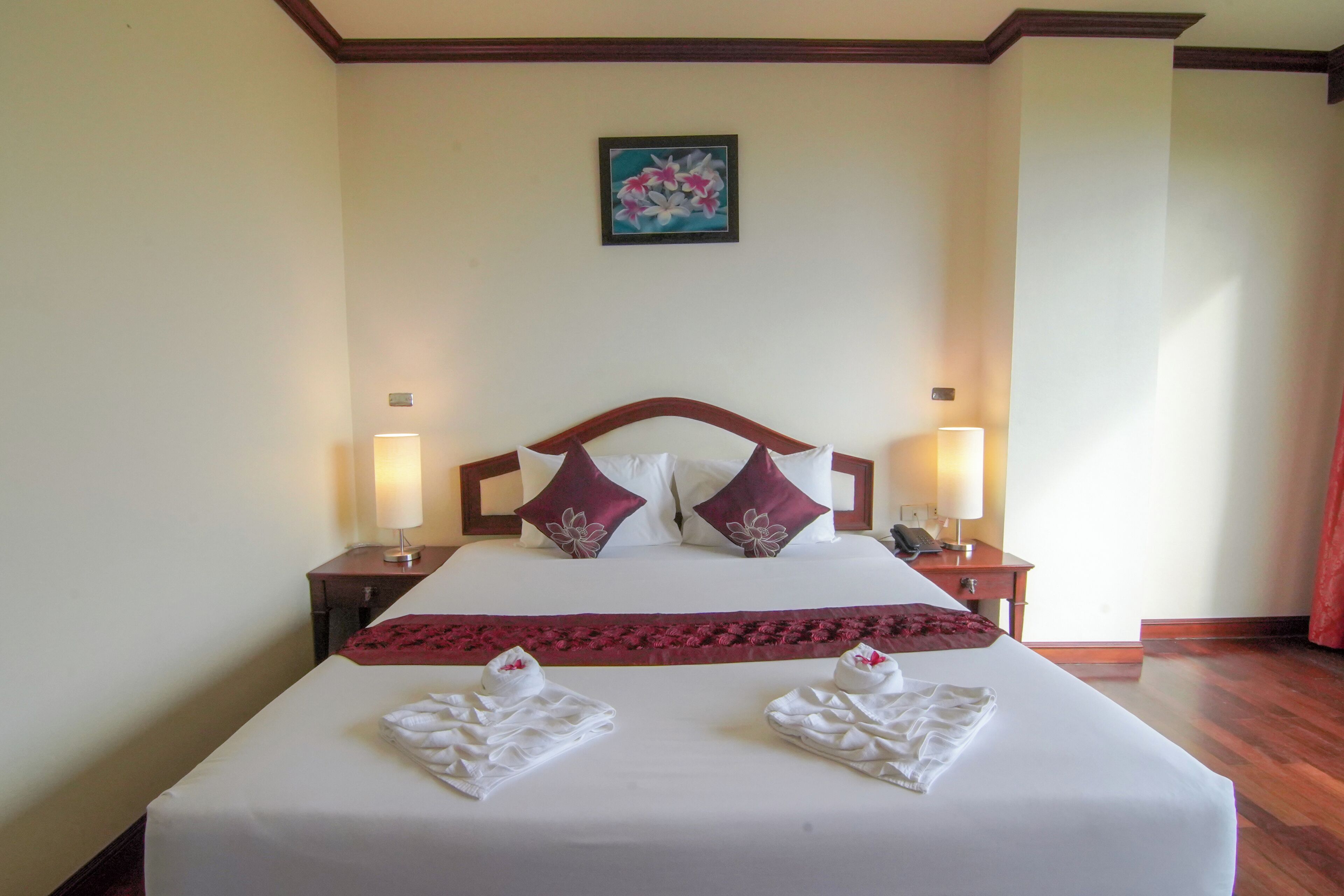 A neatly made bed with white linens and two decorative pillows, a red throw blanket, and two towels folded on the bed. There are two bedside lamps on wooden nightstands, and a framed artwork hangs on the wall above the bed.