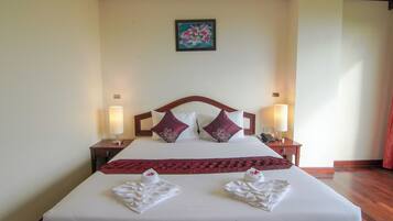 A neatly made bed with white linens and two decorative pillows, a red throw blanket, and two towels folded on the bed. There are two bedside lamps on wooden nightstands, and a framed artwork hangs on the wall above the bed.