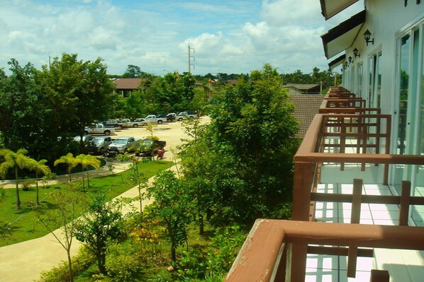 A view from a balcony overlooking a parking area with several cars, lush greenery, and a building with large windows and a balcony railing.