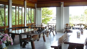 A spacious indoor dining area with wooden tables and benches, large windows with a view of greenery outside. - Thongpaeka Hotel (Prasat)