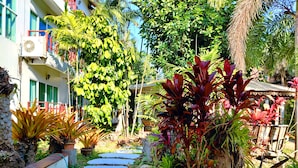 A garden with a stone pathway, various plants, and a building with a red railing and air conditioning unit. - Thongpaeka Hotel (Prasat)