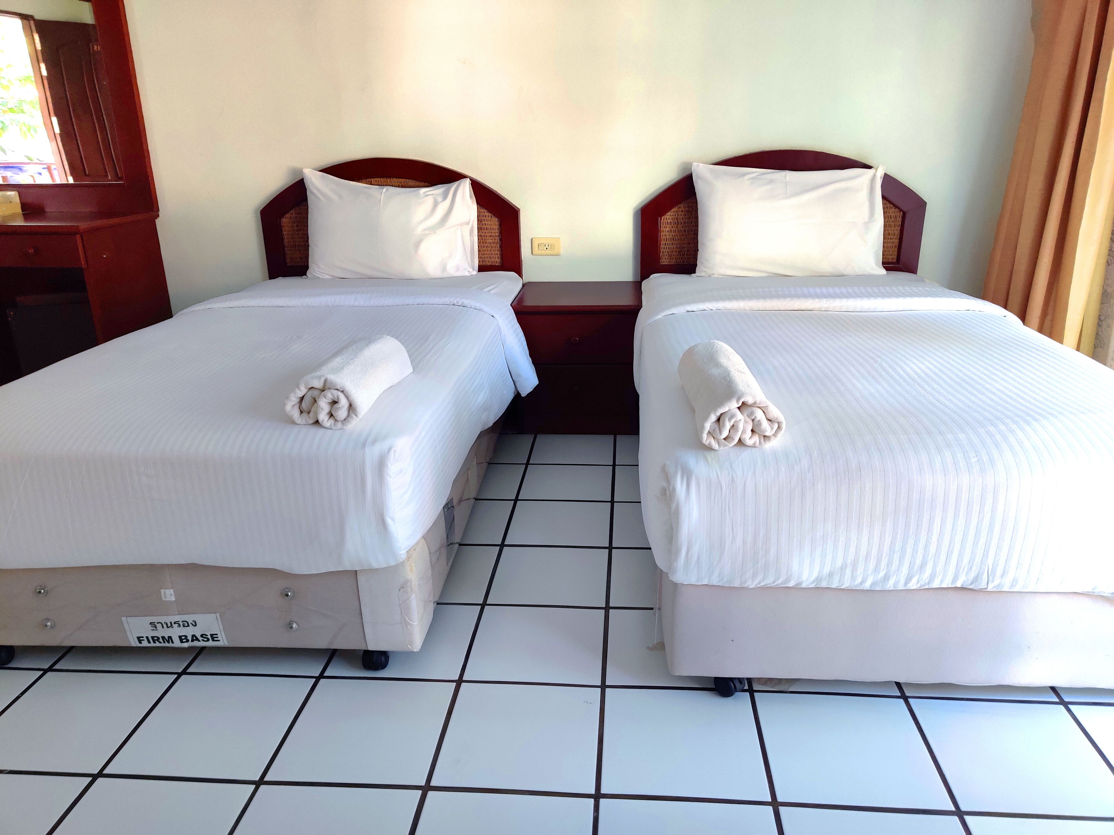 Two single beds with white linens and rolled towels, a wooden headboard, and a tiled floor.