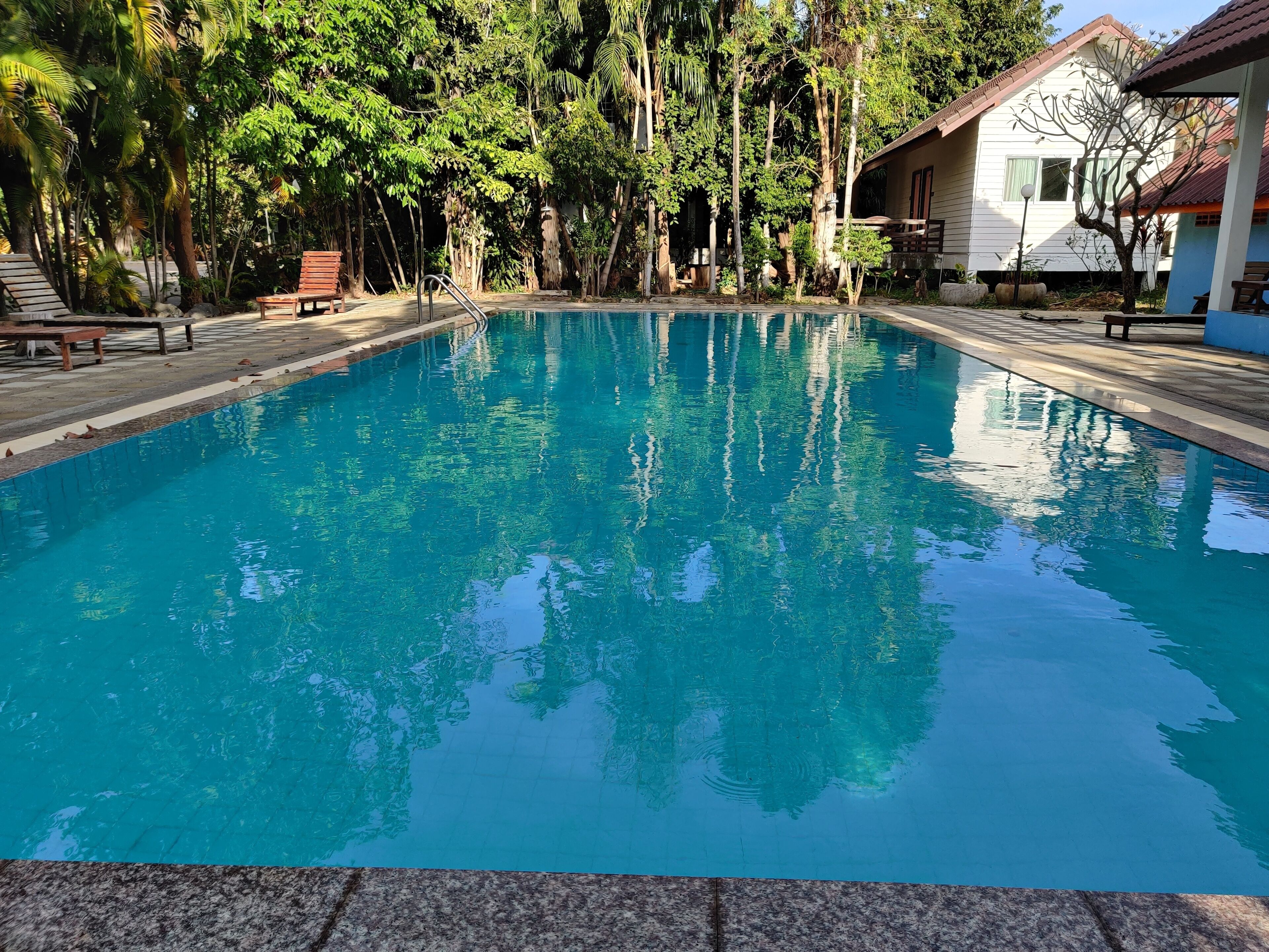 A clear swimming pool surrounded by trees and a building with a tiled roof.