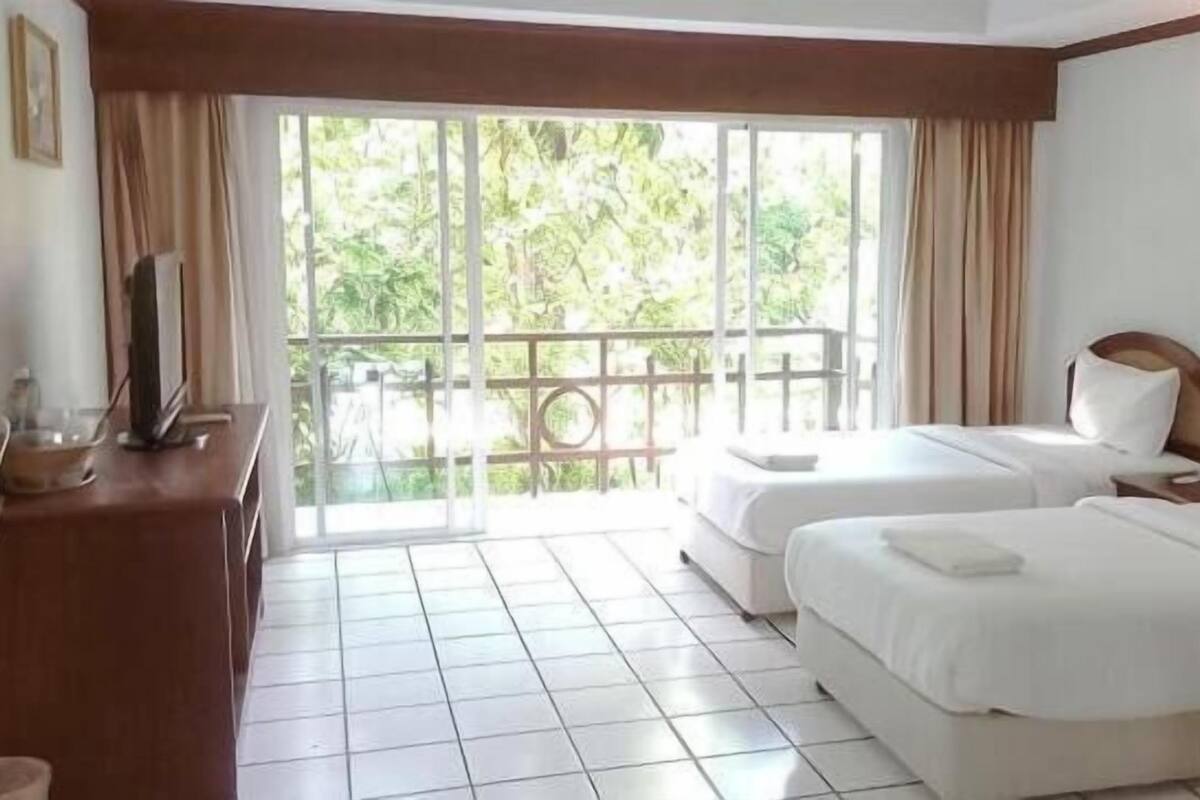A hotel room with two beds, a TV on a wooden table, and a balcony with a view of greenery.