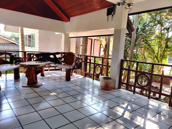 A covered patio area with a wooden bench and table, a tiled floor, and a view of greenery outside.