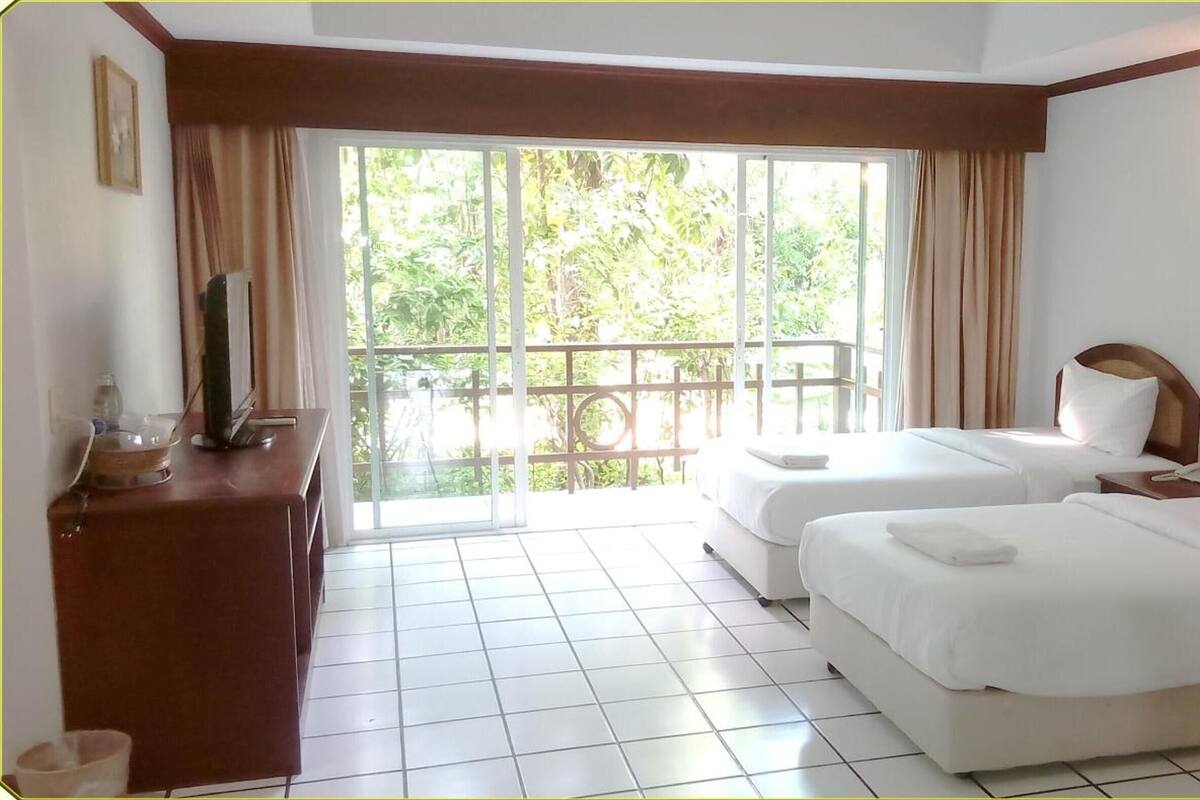 A hotel room with two beds, a desk, a television, and a large window with a view of greenery.