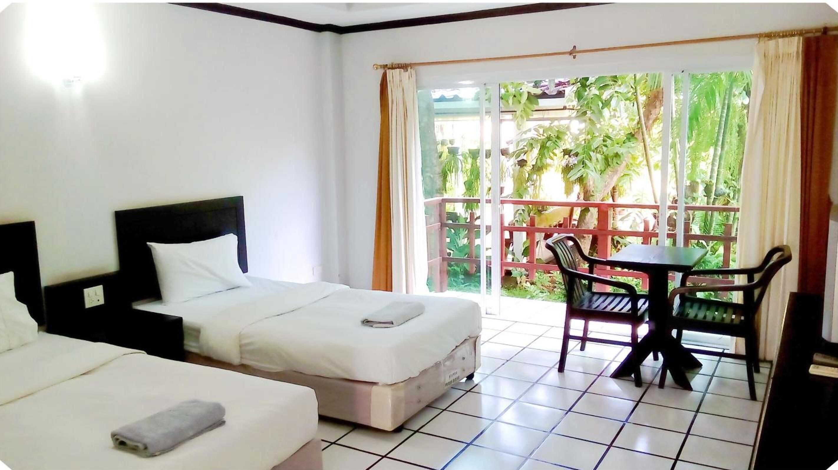 A hotel room with two beds, a small table, and a balcony with a view of greenery.