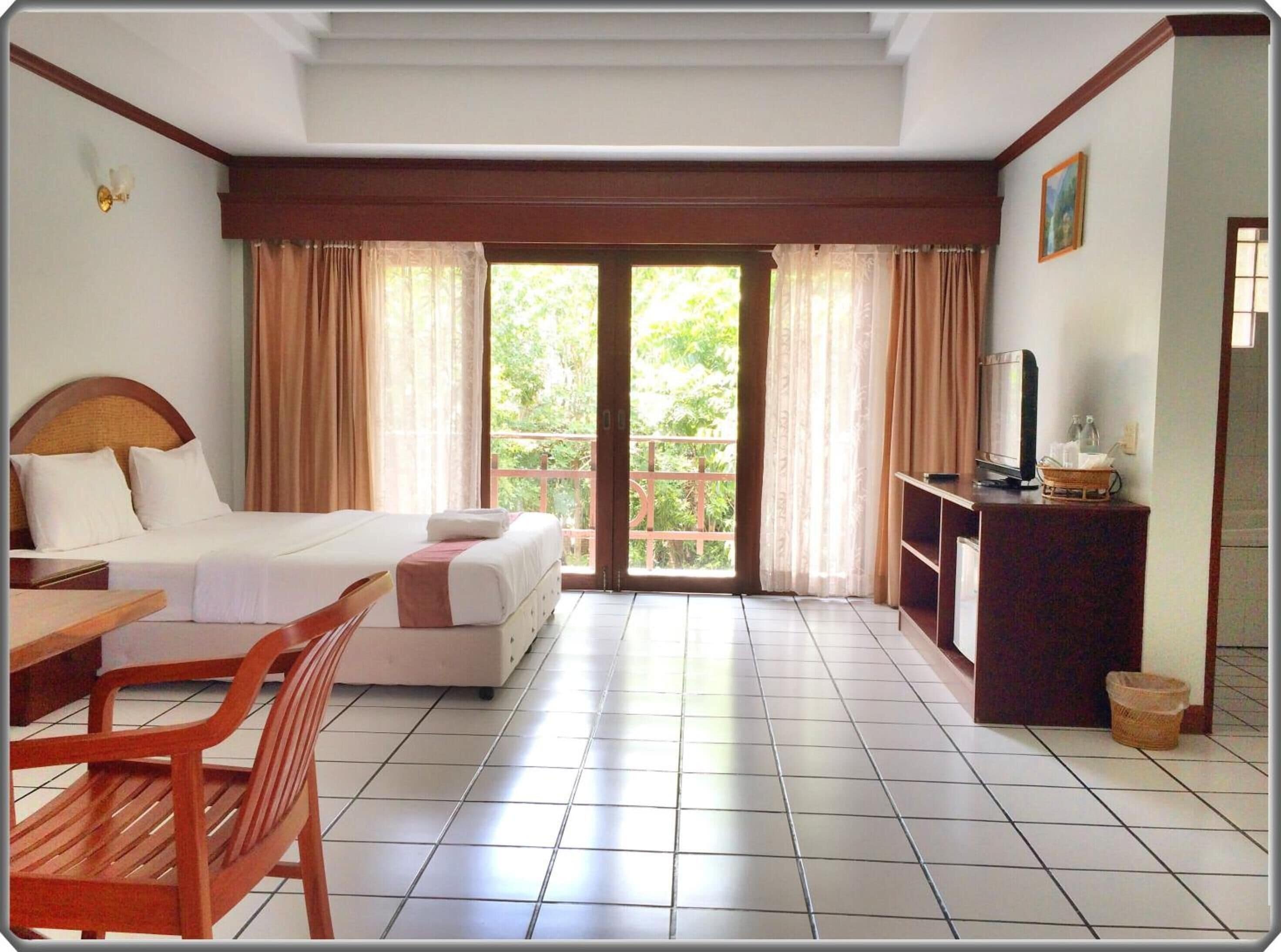 Una camera d'hotel con un letto, una scrivania, una sedia, una televisione e un balcone con tende.