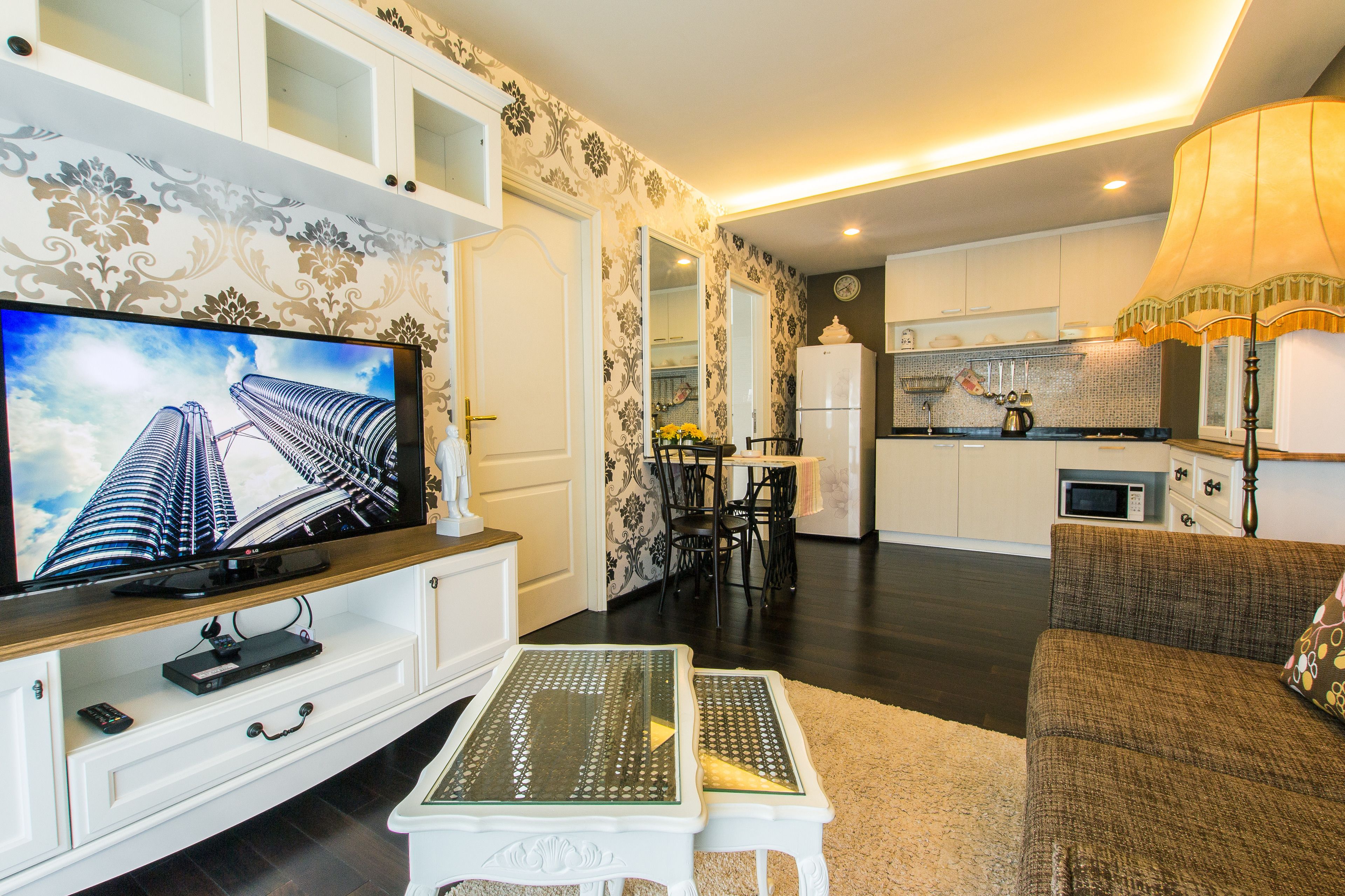 A modern living room with a flat-screen TV, a dining area with chairs, a kitchen with appliances, and a sofa with a lamp.