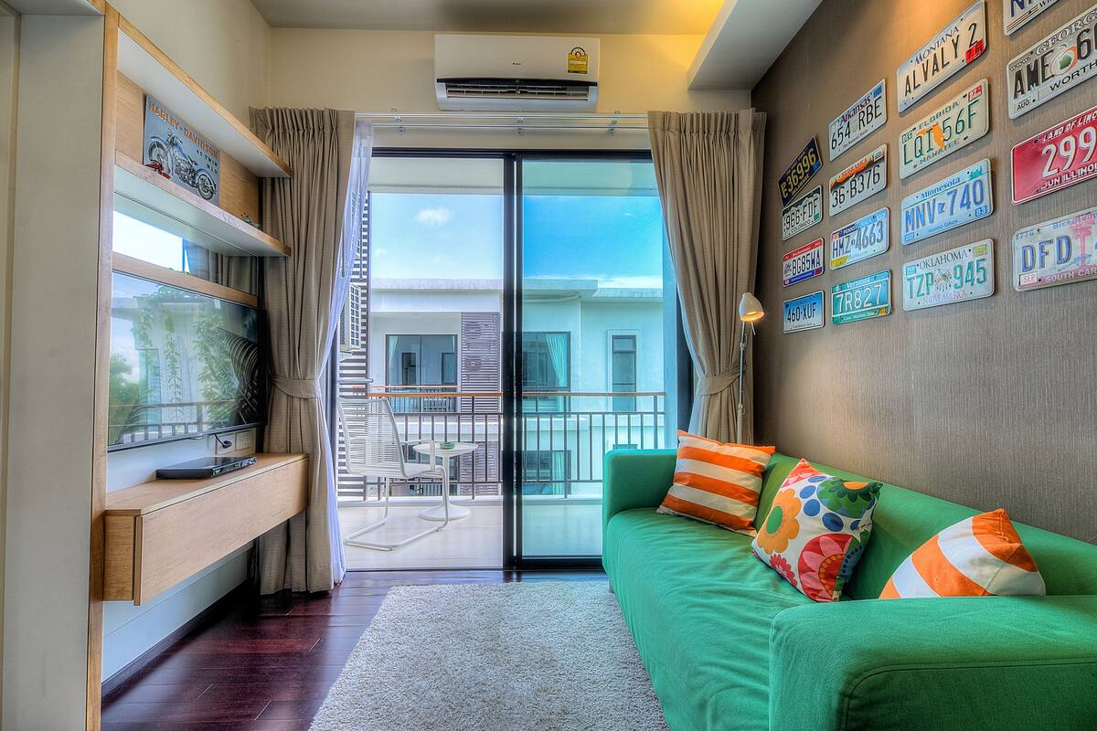 A modern living room with a green sofa, a TV on a wooden shelf, and a balcony with a view of buildings.