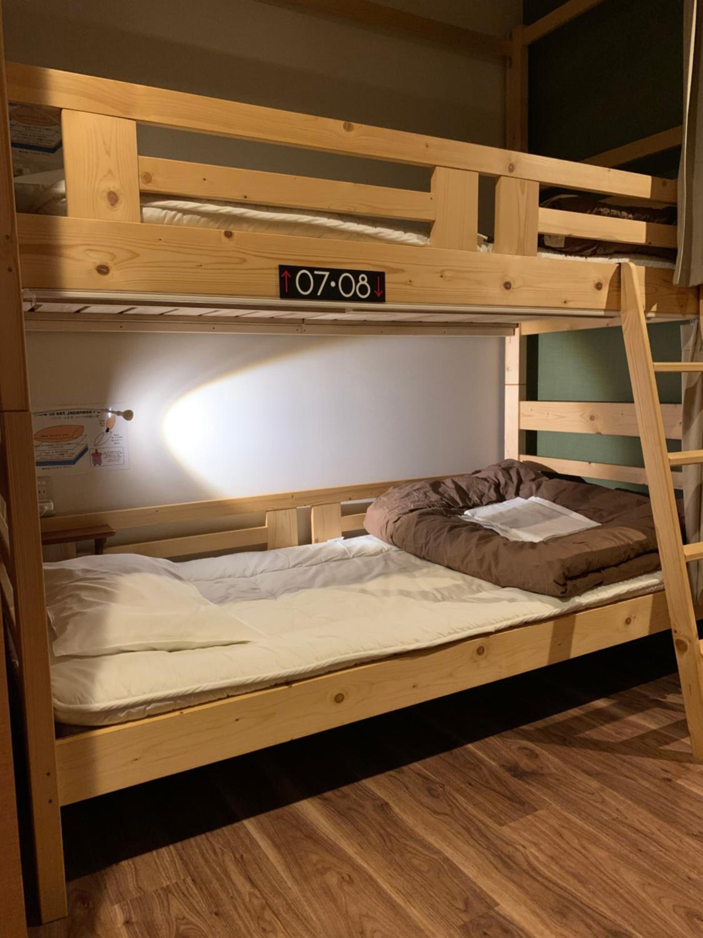 Una cama litera con estructura de madera, colchón blanco y almohada marrón. Cuenta con una escalera unida al costado.