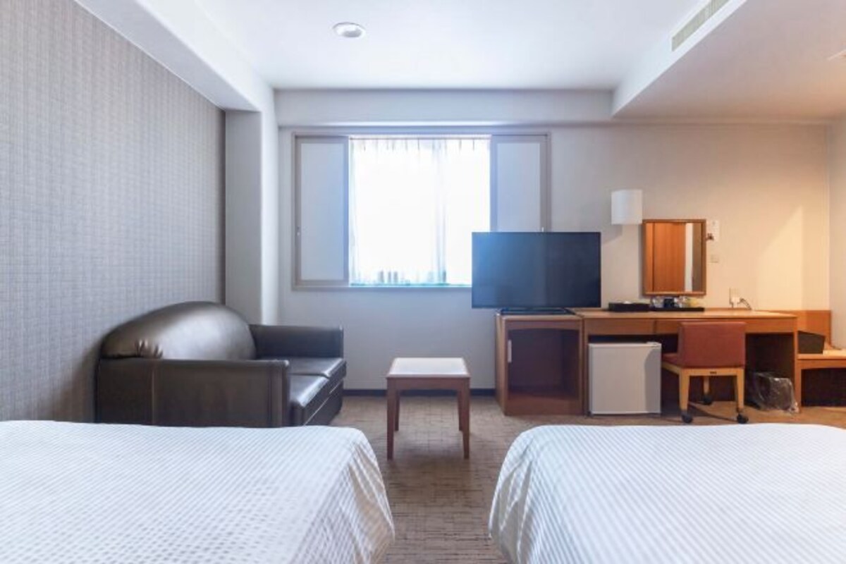 Habitación de hotel con dos camas, un escritorio, televisión, una mesita y una ventana con cortinas.