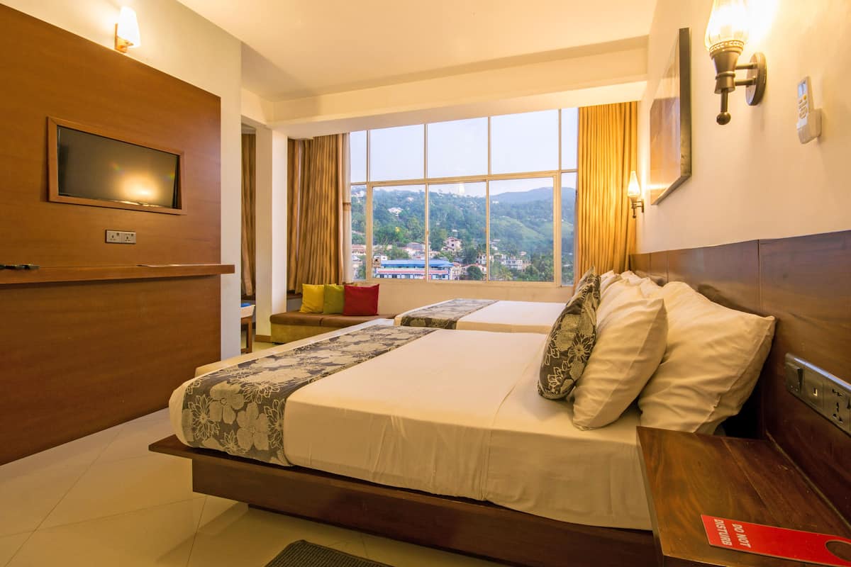 A hotel room with a large bed, a TV mounted on the wall, a window with a view of the hills, and a bedside table with a red sign.