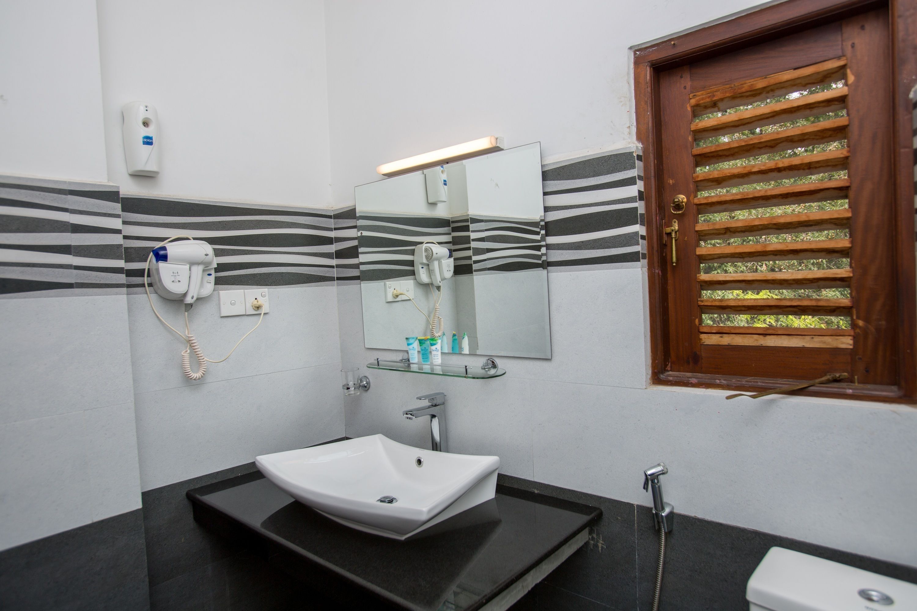 A bathroom with a white sink, a mirror, a towel dispenser, a hairdryer, and a window with blinds.