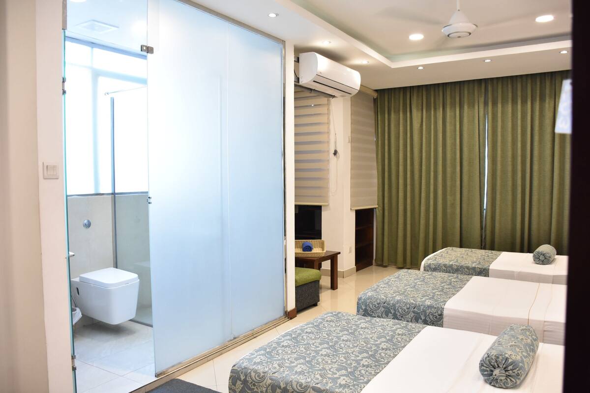 A hotel room with a bathroom, two beds, a small table, and a green curtain.