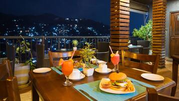 Une table dressée pour le dîner, avec un hamburger, des frites et des boissons, donnant sur une vue panoramique de la ville la nuit.