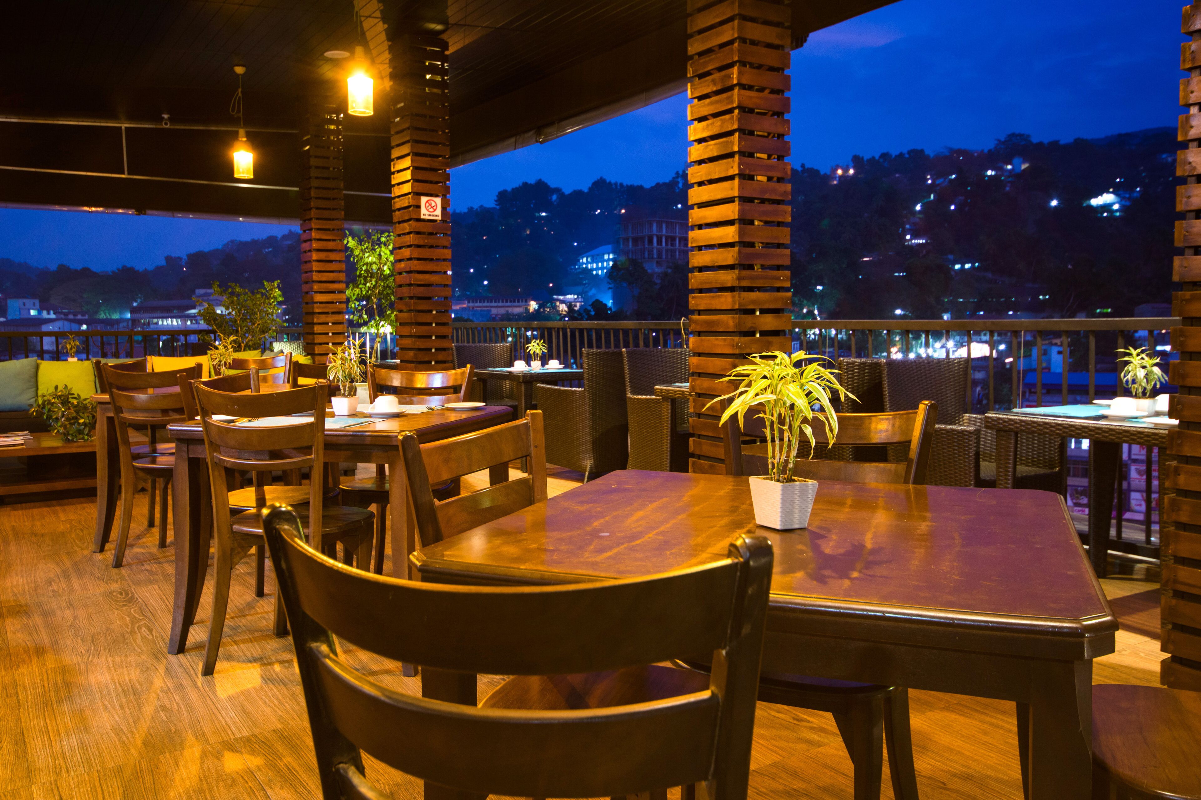 An outdoor dining area with wooden tables and chairs, potted plants, and a view of a cityscape at dusk.